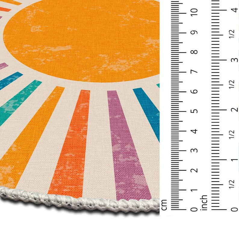 Op de afbeelding: Een rond, wit en oranje kleed met een zonontwerp. De zon is geel met oranje stralen. De stralen zijn omrand met verschillende kleuren, waaronder blauw, groen, paars en oranje.