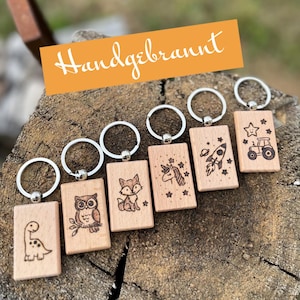 Puede incluir: Una colección de llaveros de madera con anillas plateadas. Cada llavero rectangular presenta un diseño grabado diferente: un dinosaurio, un búho, un zorro, un unicornio, un cohete y un tractor. Los llaveros se muestran sobre una superficie de madera, con la palabra "Handgebrannt" en un rectángulo naranja.