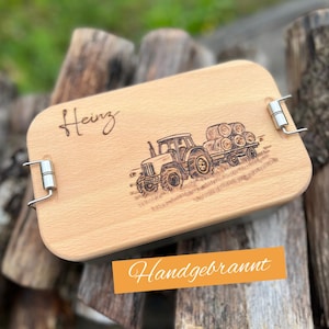 Könnte beinhalten: Ein helles Holzserviertablett mit Metallgriffen. Das Tablett zeigt einen gravierten Traktor, der einen Heuballenanhänger zieht, und den Namen "Heinz" in Schreibschrift. Der Text "Handgebrannt" befindet sich auf einem orangefarbenen Etikett.