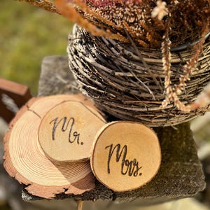 Pode incluir: Duas fatias redondas de madeira com as palavras "Mr." e "Mrs." gravadas na superfície. As fatias estão sobre uma superfície de madeira.