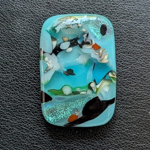 May include: A rectangular glass cabochon featuring a light blue base with embedded abstract patterns in black, white, and orange. The surface includes iridescent, shimmering accents and smooth, rounded edges, creating a multi-layered appearance.