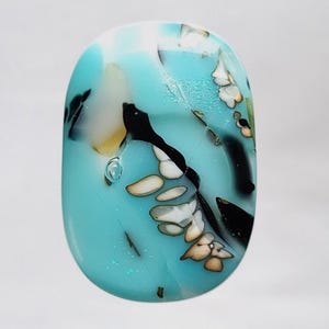 May include: An oval-shaped gemstone with a light blue base, featuring black, white, and tan organic patterns. The stone has a polished, glossy finish, with a few small bubbles visible within the translucent material. The design resembles a stylized landscape.