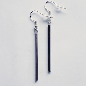 May include: A pair of silver-toned drop earrings featuring long, slender, rectangular bar pendants. Each earring is attached to a curved ear wire hook with a small metal bead detail and includes a clear silicone backing for secure placement.