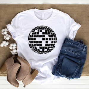 May include: White t-shirt with a black disco ball graphic. The shirt is laying on a brown burlap surface with a pair of brown suede ankle boots and blue jeans.