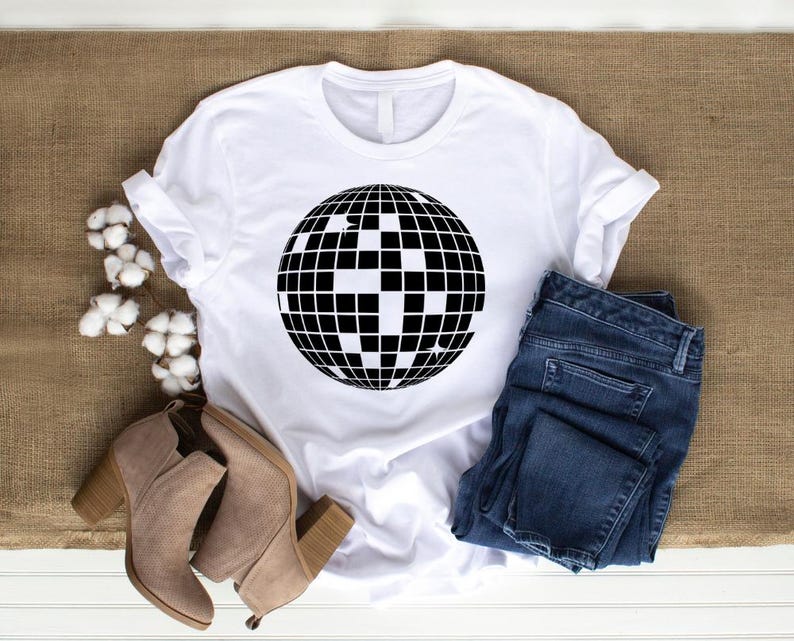May include: White t-shirt with a black disco ball graphic. The shirt is laying on a brown burlap surface with a pair of brown suede ankle boots and blue jeans.