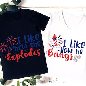 May include: Two t-shirts, one navy blue and one white, with patriotic designs. The navy shirt says "I Like How She Explodes" with fireworks. The white shirt says "I Like How He Bangs" with a firework rocket.