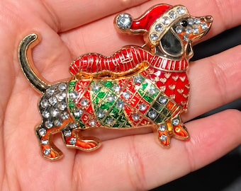 May include: A gold-tone dachshund brooch featuring red and green enamel detailing in a diamond pattern, accented with clear rhinestones throughout the body. The dog wears a red enamel Santa hat with a rhinestone trim and a red sweater.