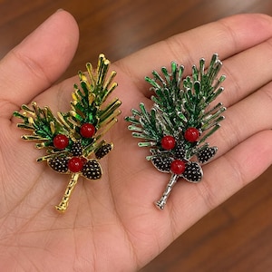 May include: Two pine branch brooches, one with a gold-tone finish and one with a silver-tone finish. Both feature green enamelled needles, three small red spherical accents, and dark brown textured pinecone details at the base of the branches.