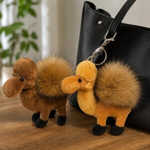 May include: Two plush camel keychains with soft brown and tan fur, black hooves, and fluffy faux fur humps. One camel is attached to a silver metal lobster clasp and keyring, which is clipped onto a large black leather tote bag.