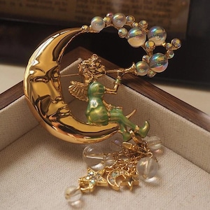 May include: A gold-toned brooch featuring a crescent moon with a face, a green-clad figure playing a trumpet, and iridescent spherical accents. Dangling from the base are gold chains with small star and moon charms and clear beads.