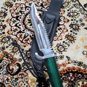 May include: A survival knife with a serrated blade and a green wrapped handle. The knife is in a black leather sheath with a snap closure. The blade is silver and the handle is green. The knife is designed for outdoor use.