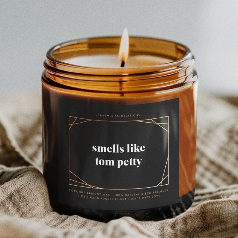 Pu&ograve; includere: Una candela di vetro marrone con un'etichetta nera che recita "smells like tom petty". L'etichetta ha un bordo dorato e il testo &egrave; bianco. La candela &egrave; accesa e la fiamma &egrave; visibile.