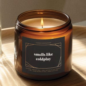 May include: A brown glass candle jar with a black label that reads "CHARMED SCENTSATIONS" and "smells like coldplay". The candle is lit and the wax is melting. The label also includes the text "COCONUT APRICOT WAX | 100% NATURAL & ECO FRIENDLY | 4 OZ | HAND POURED IN USA | MADE WITH LOVE."