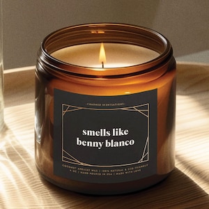 May include: A brown glass candle jar with a black label that reads "smells like benny blanco". The label also includes the text "CHARMED SCENTSATIONS", "COCONUT APRICOT WAX", "100% NATURAL & ECO FRIENDLY", "4 OZ | HAND POURED IN USA | MADE WITH LOVE".