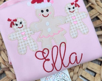 May include: Pink long-sleeved shirt with three gingerbread people appliqués. The center gingerbread person wears a Santa hat. The name "Ella" is embroidered in red script. A tag with the text "Sweet Stitches embroidery" is visible.