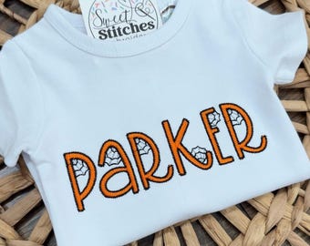 May include: White short-sleeved baby shirt with the name "PARKER" embroidered in orange thread. The letters are outlined in black and feature spiderweb designs. A small tag with the text "Sweet Stitches" is attached.