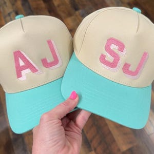 May include: Two baseball caps with cream-colored crowns and teal brims are held in a hand. The caps have pink embroidered initials, one with "AJ" and the other with "SJ". The caps have a teal button on top.