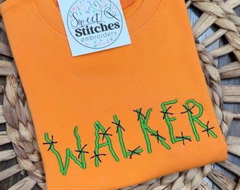 May include: Orange t-shirt with the name "WALKER" embroidered in green thread with black cross-stitch details. A white tag with the text "Sweet Stitches embroidery" is attached to the shirt.
