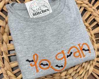 May include: A gray t-shirt with the name "Logan" embroidered in orange, with small black bat designs. The shirt is folded and rests on a woven, brown surface. A white tag with the text "Sweet Stitches embroidery" is visible.