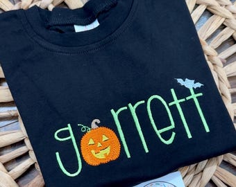 May include: Black t-shirt with the name "Garrett" embroidered in green, featuring an orange jack-o'-lantern and a bat. The shirt is displayed on a woven surface, with a "Sweet Stitches Embroidery" sticker.