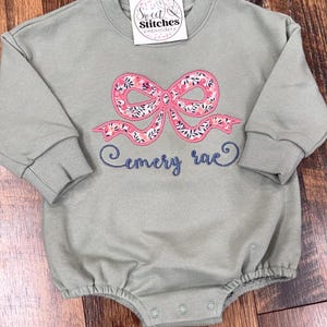 May include: A sage green long-sleeved baby onesie with a pink floral bow and the name "emery rae" embroidered in blue thread. The onesie has a round neckline and snaps at the bottom.