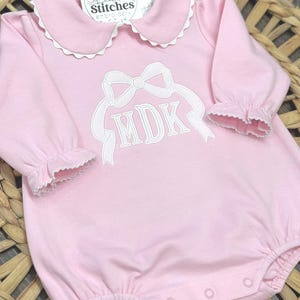 May include: A light pink baby romper with a white Peter Pan collar and ruffled cuffs. The romper features a white embroidered bow and the initials "MDK". The tag reads "Sweet Stitches Embroidery".