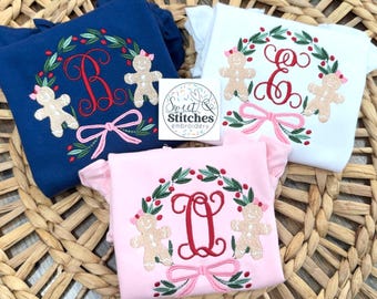 May include: Three shirts with embroidered Christmas designs. Each shirt features a wreath with a letter, gingerbread men, and a pink bow. The shirts are navy blue, white, and pink.