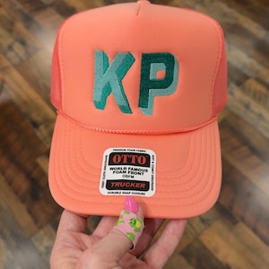 May include: Coral-colored trucker hat with a mesh back and the embroidered letters "KP" in teal. The hat features a curved brim and a snap closure. The hat has a label that says "OTTO WORLD FAMOUS FOAM FRONT OSFM TRUCKER DURABLE SNAP CLOSURE".