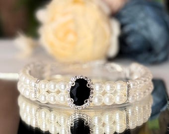 May include: A double-strand pearl bracelet with a black rhinestone center piece. The bracelet is silver and has a delicate design.