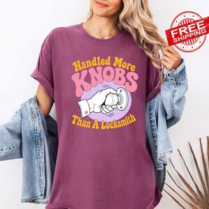 May include: A plum-coloured short-sleeve t-shirt featuring a graphic print of a hand turning a doorknob. The text reads Handled More KNOBS Than A Locksmith in yellow and pink lettering. The shirt is worn with a light blue denim jacket.