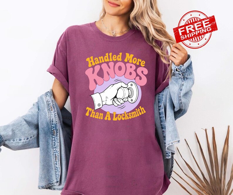 May include: A plum-coloured short-sleeve t-shirt featuring a graphic print of a hand turning a doorknob. The text reads Handled More KNOBS Than A Locksmith in yellow and pink lettering. The shirt is worn with a light blue denim jacket.