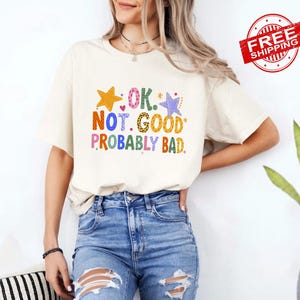May include: A cream-colored short-sleeve t-shirt featuring the colorful text OK. NOT. GOOD. PROBABLY BAD. The letters are filled with various patterns including leopard print, polka dots, and checkers, accompanied by yellow and purple star graphics.