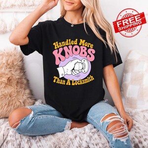 May include: A black short-sleeved t-shirt featuring a graphic print of a hand turning a round doorknob. The text reads Handled More KNOBS Than A Locksmith in yellow and pink lettering. The design is centred on the front of the garment.