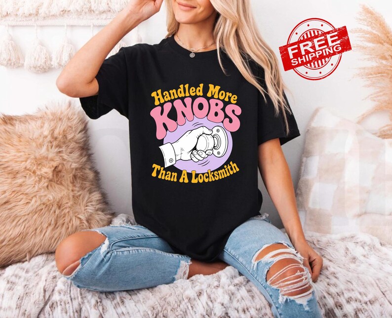 May include: A black short-sleeved t-shirt featuring a graphic print of a hand turning a round doorknob. The text reads Handled More KNOBS Than A Locksmith in yellow and pink lettering. The design is centred on the front of the garment.