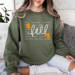 May include: An olive green sweatshirt with the word "fall" in white script. The word is surrounded by a list of fall-themed words and phrases, including "sweaters," "leaves," and "harvest." Two orange leaves are on the right and left of the word "fall."