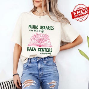 May include: A cream-colored t-shirt featuring the dark green text "PUBLIC LIBRARIES are the only DATA CENTERS i support" with a central graphic of an open book with pink floral illustrations emerging from the pages, paired with blue denim jeans.