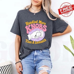 May include: A dark charcoal grey short-sleeve t-shirt featuring a graphic print of a hand turning a doorknob. The text reads Handled More KNOBS Than A Locksmith in yellow and pink retro-style lettering centred on the chest area.