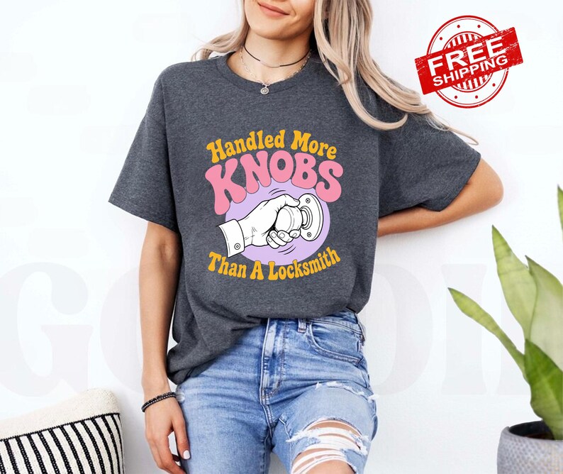 May include: A dark charcoal grey short-sleeve t-shirt featuring a graphic print of a hand turning a doorknob. The text reads Handled More KNOBS Than A Locksmith in yellow and pink retro-style lettering centred on the chest area.