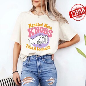 May include: A cream-colored short-sleeve t-shirt featuring a graphic of a hand turning a doorknob. The text reads Handled More KNOBS Than A Locksmith in orange and pink lettering. The design is centered on the chest of the garment.