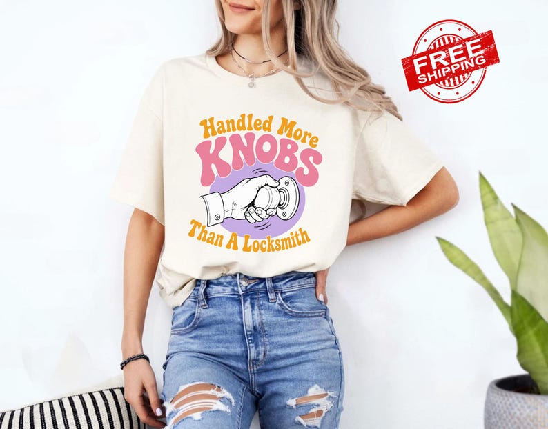May include: A cream-coloured short-sleeve t-shirt featuring a graphic of a hand turning a doorknob. The text reads Handled More KNOBS Than A Locksmith in orange and pink lettering. The design is centred on the chest of the garment.