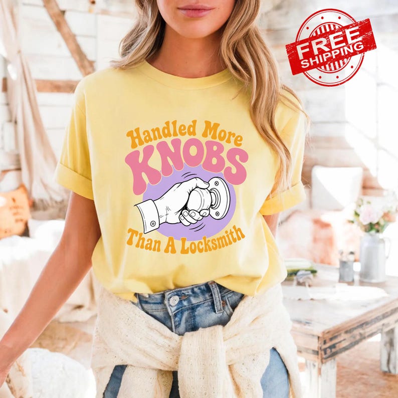 May include: A pale yellow short-sleeve t-shirt featuring a graphic of a hand turning a doorknob. The text reads Handled More KNOBS Than A Locksmith in orange and pink lettering. The shirt is paired with blue denim jeans and a white sweater.