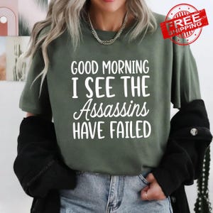 May include: An olive green short-sleeved t-shirt featuring white text that reads GOOD MORNING I SEE THE ASSASSINS HAVE FAILED. The shirt is worn with a black jacket and blue denim jeans, and a silver chain necklace is visible around the neck.
