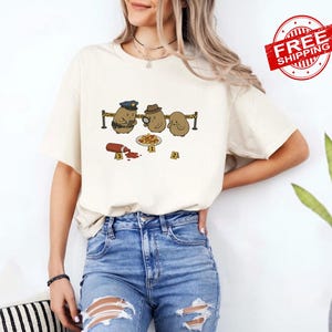 May include: A cream-colored short-sleeve t-shirt featuring a graphic of three cartoon potatoes at a crime scene. One potato wears a police hat, another uses a magnifying glass, and a third stands nearby. A bottle of ketchup and evidence markers are present.