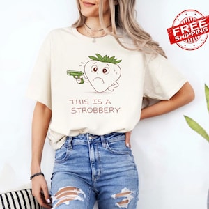 May include: Cream-colored t-shirt featuring a cartoon strawberry holding a green gun, with the text "THIS IS A STROBBERY" below. The strawberry has a worried expression. The shirt is paired with blue jeans.