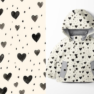 Könnte beinhalten: Ein cremefarbener Kinderregenmantel mit schwarzem Herzmuster und grauen Akzenten. Die Jacke hat eine Kapuze, Taschen und eine Knopfleiste. Das Muster umfasst schwarze Herzen und kleine schwarze Punkte.