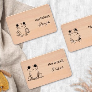Könnte beinhalten: Drei Holzschilder mit Froschillustrationen und Namen. Jedes Schild hat eine helle Holzmaserung, eine schwarze Froschzeichnung und den Text "Hier krümelt" mit einem Namen: Ronja, Dennis und Pierre. Ein kleiner Teddybär ist im Hintergrund.