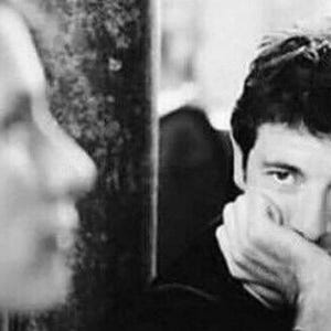 May include: A black and white photo of a man with dark hair looking down and resting his chin on his hand. He is wearing a dark shirt. The photo is out of focus in the background.