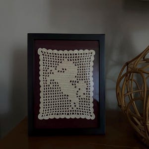 May include: A rectangular black frame containing a cream-colored crochet piece with a scalloped edge. The crochet features a central silhouette of a rearing horse created through a grid-like pattern, set against a dark maroon background.
