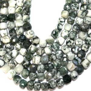 May include: Close-up of a collection of square-cut beads. The beads are a mix of white and various shades of green, with a speckled pattern. The beads are arranged closely together, suggesting they are strung together for jewelry making.