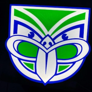 May include: A stylized emblem featuring a symmetrical design with green, white, and blue elements. The emblem has a shield-like shape with curved lines and a central, tongue-like shape. The background is black.